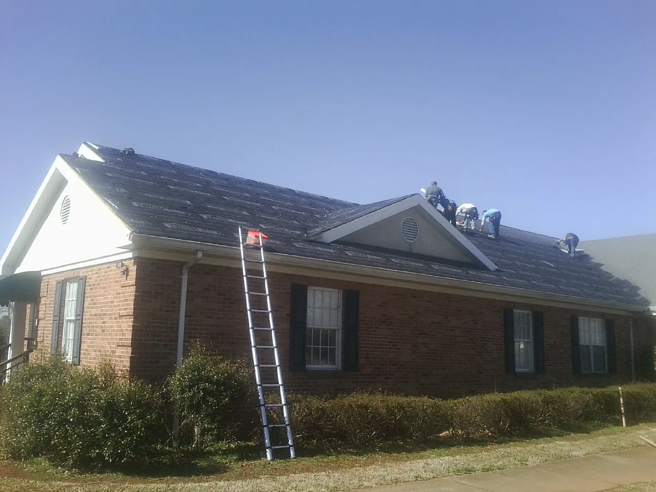 Crew tearing shingles off a brick ranch roof with a ladder set — Emergency Roof Repair work by our crew in Capac