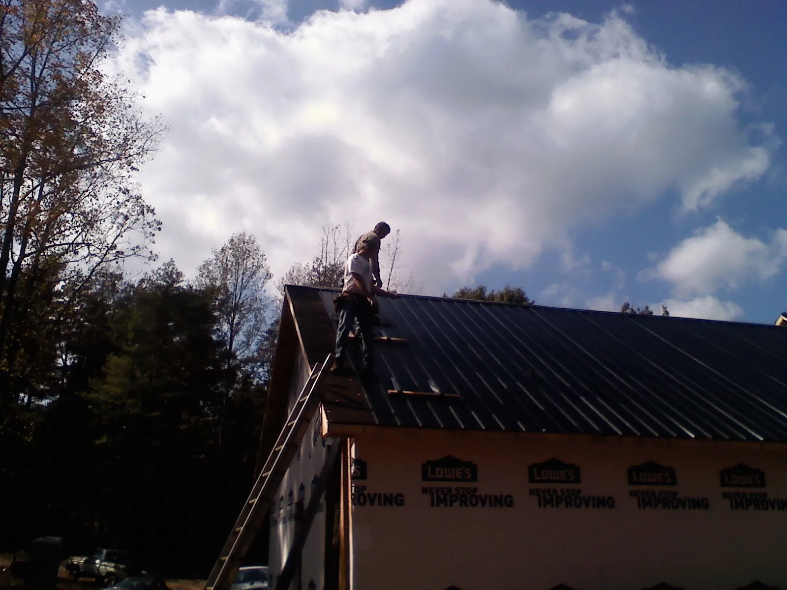 Roofer fastening dark metal panels high against a cloudy sky, from a Roof Inspection job in Capac